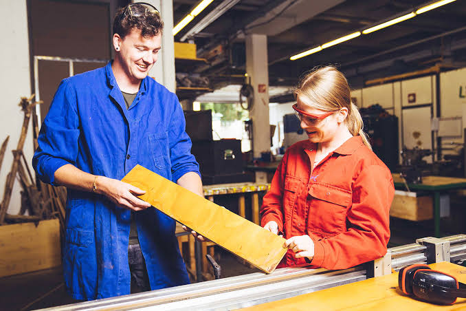 Zwei junge Menschen in blauer und roter Arbeitskleidung prüfen gemeinsam ein Werkstück in einer Werkstatt.